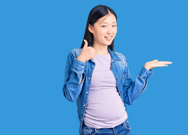Молодая красивая китаянка, беременная, ждет ребенка, показывающего пальмовую руку и делающего все хорошо, жест с большим пальцем вверх, улыбаясь счастливой и веселой  - Фото, изображение