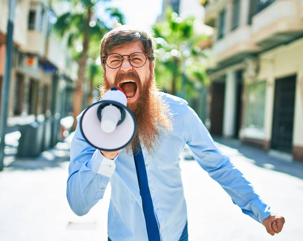 Young redhead businessman with angry expression. Screaming using megaphone at street of city. - Photo, Image
