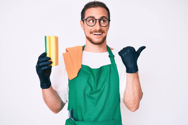 Young handsome man wearing apron holding scourer pointing thumb up to the side smiling happy with open mouth  - Photo, Image