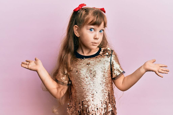 Маленька кавказька дівчинка, яка носить святкові блискітки, одягнена в безглузді і заплутані вирази з піднятими руками. концепція сумніву
.  - Фото, зображення