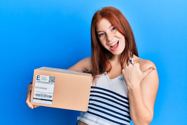 Joven cabeza roja chica sosteniendo paquete de entrega apuntando pulgar hacia el lado sonriendo feliz con la boca abierta  - Foto, Imagen