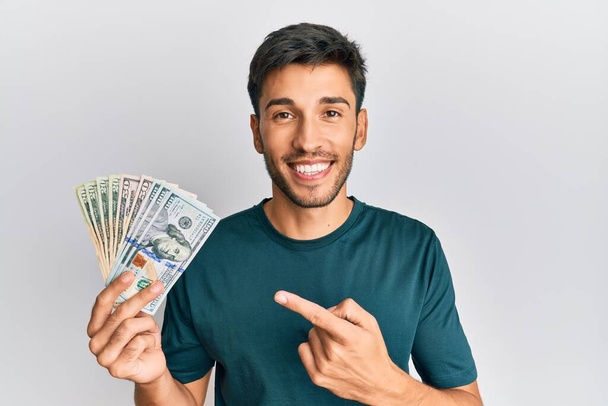 Młody przystojny mężczyzna trzyma dolary uśmiechając się szczęśliwy wskazując ręką i palcem  - Zdjęcie, obraz