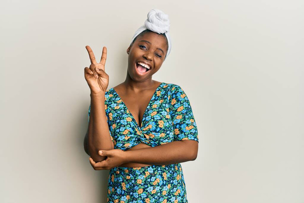 Junge Afrikanerin mit Haarturban und afrikanischem Stil lächelt mit glücklichem Gesicht und zwinkert in die Kamera, als sie ein Siegeszeichen macht. Nummer zwei.  - Foto, Bild