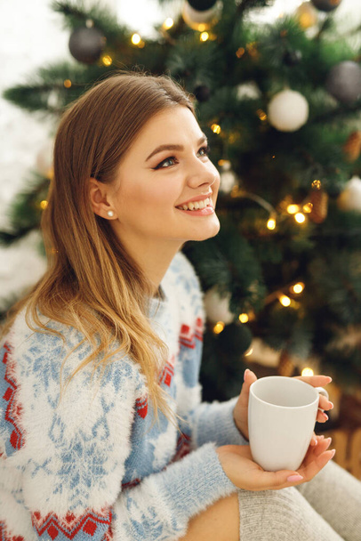 Close up of girl wears festive clothes take cup of coffee at home near Christmas Tree. Smiling woman sit on mat in blue sweater with deer and gray stockings - Zdjęcie, obraz