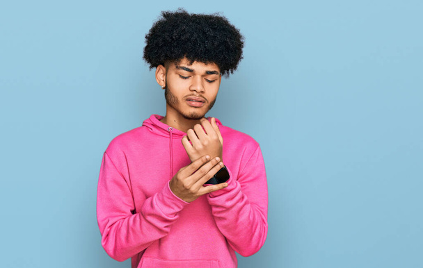 Jeune homme afro-américain aux cheveux afro portant un sweat rose décontracté souffrant de douleur aux mains et aux doigts, inflammation de l'arthrite  - Photo, image