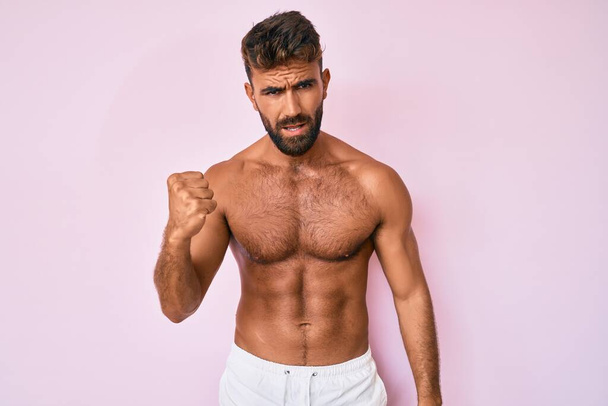 Young hispanic man standing shirtless angry and mad raising fist frustrated and furious while shouting with anger. rage and aggressive concept.  - Photo, Image