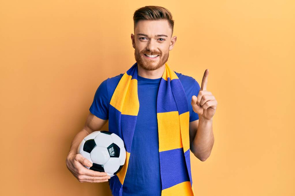 Fiatal vörös férfi futball huligán kezében labda mosolyogva egy ötlettel vagy kérdéssel ujjal mutogatva boldog arc, első számú  - Fotó, kép