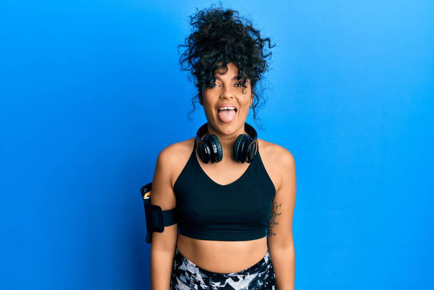 Young hispanic woman wearing gym clothes and using headphones sticking tongue out happy with funny expression. emotion concept.  - Foto, immagini