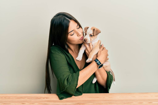 Junges hispanisches Mädchen umarmt und küsst Hund, der auf dem Tisch vor isoliertem weißen Hintergrund sitzt. - Foto, Bild