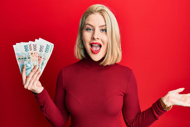 Молодая блондинка с банкнотами чех коруна празднует достижение с счастливой улыбкой и выражение победителя с поднятой рукой  - Фото, изображение