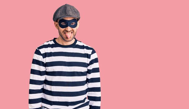 Young handsome man wearing burglar mask sticking tongue out happy with funny expression. emotion concept.  - Photo, Image