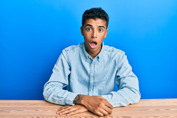 Young handsome african american man wearing casual clothes sitting on the table in shock face, looking skeptical and sarcastic, surprised with open mouth  - Foto, Imagem