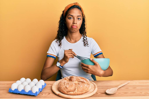 Genç Afro-Amerikalı kız masada oturmuş sıkıntılı ve endişeli, sinirli ve korkmuş bir şekilde ev yapımı ekmek yapıyor. hüzünlü ifade.  - Fotoğraf, Görsel