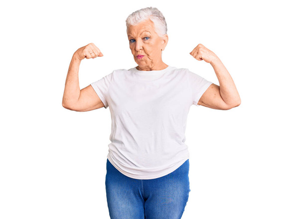 Senior bella donna con gli occhi azzurri e capelli grigi indossa casual maglietta bianca che mostra i muscoli delle braccia sorridente orgoglioso. concetto di fitness.  - Foto, immagini