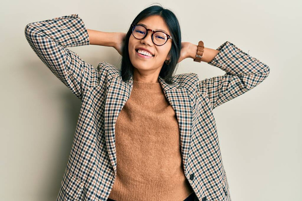 Fiatal kínai nő visel üzleti stílus és a szemüveg pihentető és nyújtózkodó, karok és kezek a fej és a nyak mögött mosolyogva boldog  - Fotó, kép