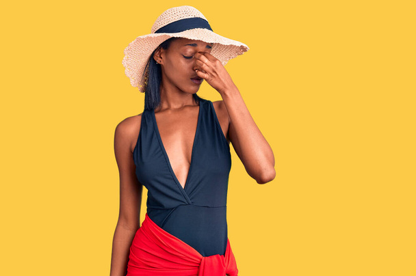 Fiatal afro-amerikai nő fürdőruhát és nyári kalapot visel fáradt dörzsölés orr és szem érzés fáradtság és fejfájás. stressz és frusztráció fogalma.  - Fotó, kép