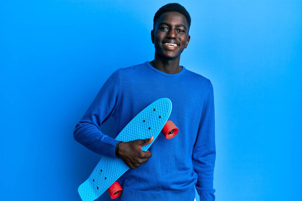 Young african american man holding skate looking positive and happy standing and smiling with a confident smile showing teeth  - Foto, Imagen