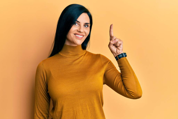 Junge kaukasische Frau in lässiger Kleidung, die mit einer Idee oder Frage lächelt und mit glücklichem Gesicht den Finger nach oben zeigt, Nummer eins  - Foto, Bild