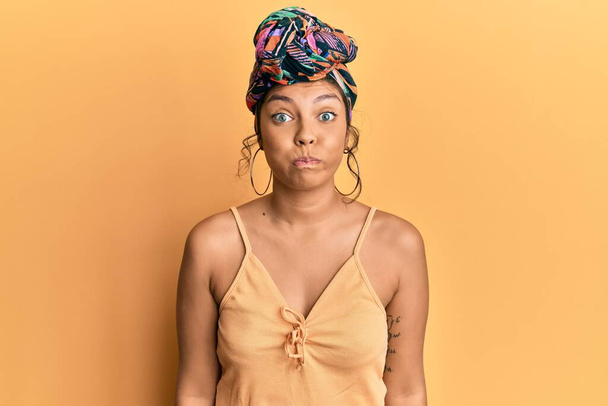 Young hispanic girl wearing hair turban over yellow background puffing cheeks with funny face. mouth inflated with air, crazy expression.  - Foto, afbeelding