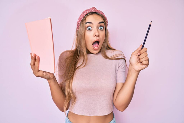 Young beautiful blonde woman holding book and pencil celebrating crazy and amazed for success with open eyes screaming excited.  - Fotoğraf, Görsel