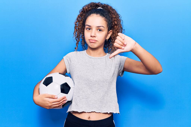 Красивая девочка с вьющимися волосами держит футбольный мяч с сердитым лицом, негативный знак показывает неприязнь с опущенными пальцами, отвержение концепции  - Фото, изображение