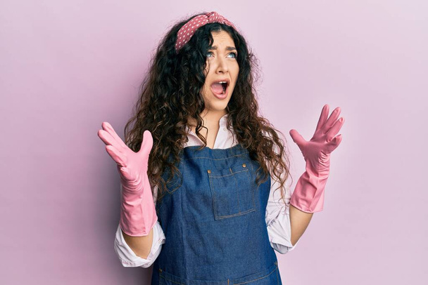Young brunette woman with curly hair wearing cleaner apron and gloves crazy and mad shouting and yelling with aggressive expression and arms raised. frustration concept.  - Photo, Image