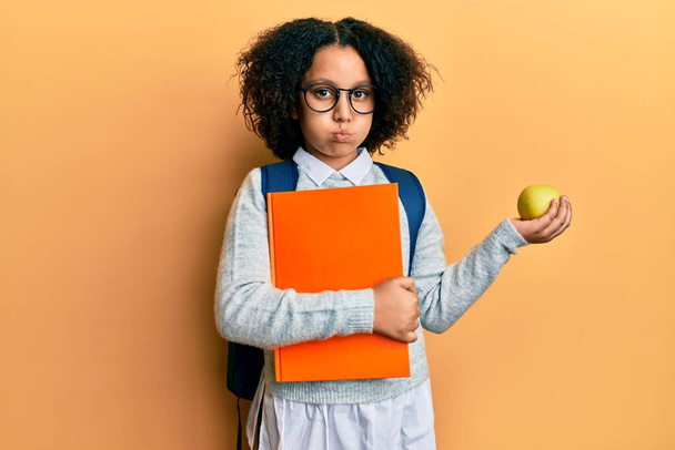Młoda dziewczynka z afro włosy noszące torbę szkolną trzymając książki i zielone jabłko puffing policzki z zabawną twarzą. usta napompowane powietrzem, łapie powietrze.  - Zdjęcie, obraz