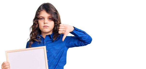 Cute hispanic child with long hair holding empty white chalkboard with angry face, negative sign showing dislike with thumbs down, rejection concept  - Photo, Image