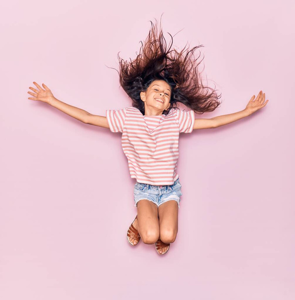 Imádnivaló spanyol kislány alkalmi ruhát visel, boldogan mosolyog. Ugrás mosollyal az arcon és a karok nyitott elszigetelt rózsaszín háttér - Fotó, kép