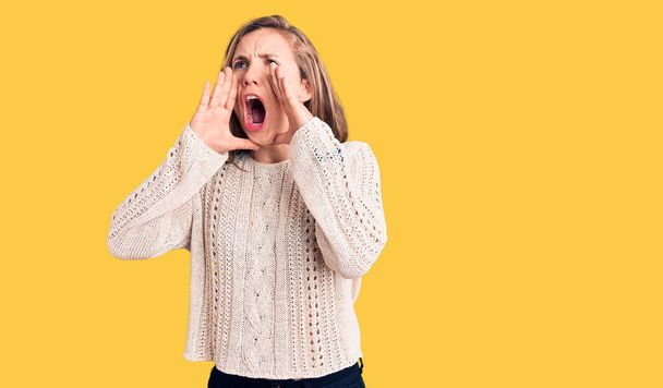 Junge schöne blonde Frau in lässigem Pullover schreit wütend laut mit den Händen über dem Mund  - Foto, Bild