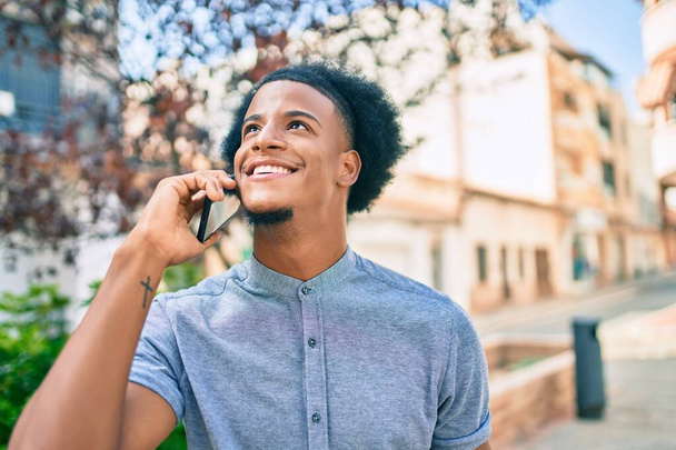 Joven hombre afroamericano sonriendo feliz hablando en el teléfono inteligente en la ciudad. - Foto, imagen