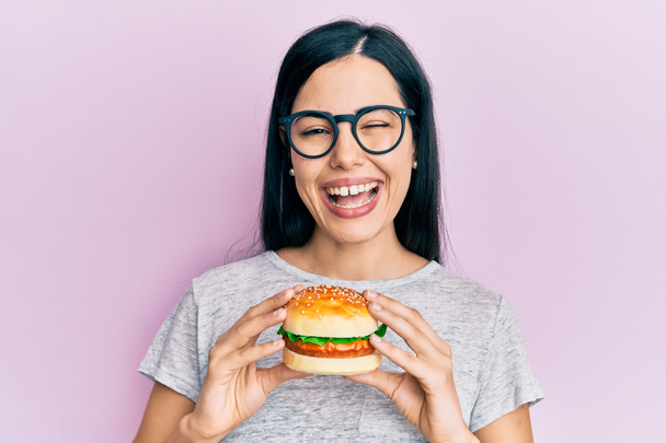 美しい若い女性が食べるおいしいハンバーガーウィンクセクシーな表情でカメラを見て,陽気で幸せな顔.  - 写真・画像
