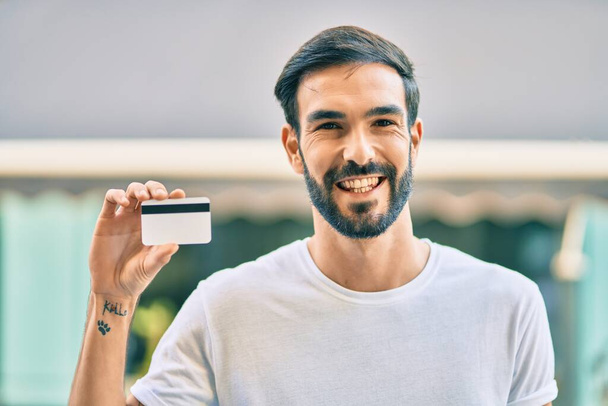 Joven hispano sonriendo feliz sosteniendo tarjeta de crédito en la ciudad. - Foto, imagen