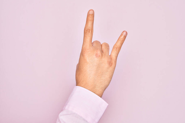 Hand van blanke jongeman met vingers over geïsoleerde roze achtergrond gebaren rock and roll symbool, met obscene hoorns gebaar - Foto, afbeelding