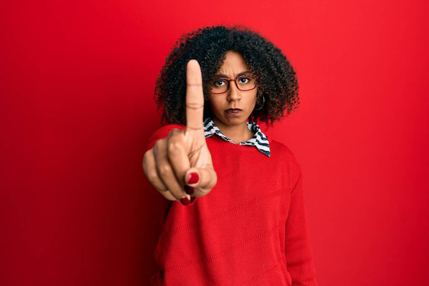Красивая африканская американка с афроволосами в свитере и очках, показывающая пальцем вверх и сердитым выражением лица, не показывающая жеста  - Фото, изображение