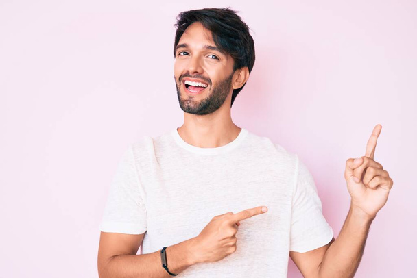Hombre hispano guapo vistiendo camiseta blanca casual sonriendo y mirando a la cámara apuntando con dos manos y dedos hacia un lado.  - Foto, Imagen