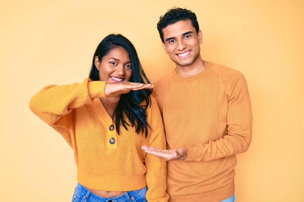 Güzel Latin genç çift, günlük giysiler giyip el kol hareketleri yaparak büyük ve büyük bir işaret, ölçü sembolü gösteriyorlar. Kameraya bakıp gülümsüyor. ölçüm kavramı.  - Fotoğraf, Görsel