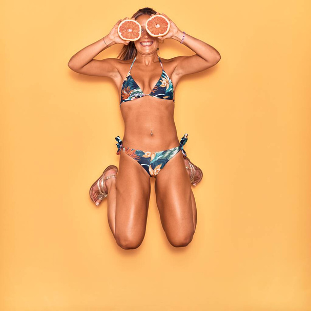 Joven mujer hispana hermosa vistiendo bikini sonriendo feliz. Saltando con sonrisa en la cara sosteniendo pomelo sobre un fondo amarillo aislado. - Foto, Imagen