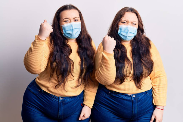 Young plus size twins wearing medical mask angry and mad raising fist frustrated and furious while shouting with anger. rage and aggressive concept.  - Photo, Image