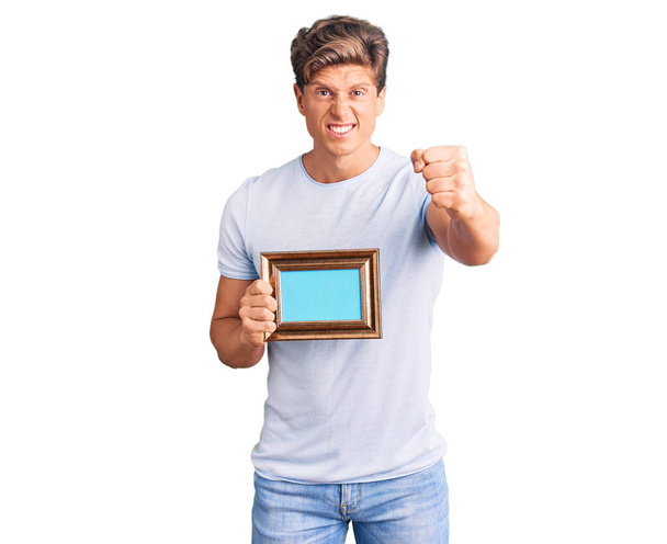 Young handsome man holding empty frame annoyed and frustrated shouting with anger, yelling crazy with anger and hand raised  - Foto, Bild