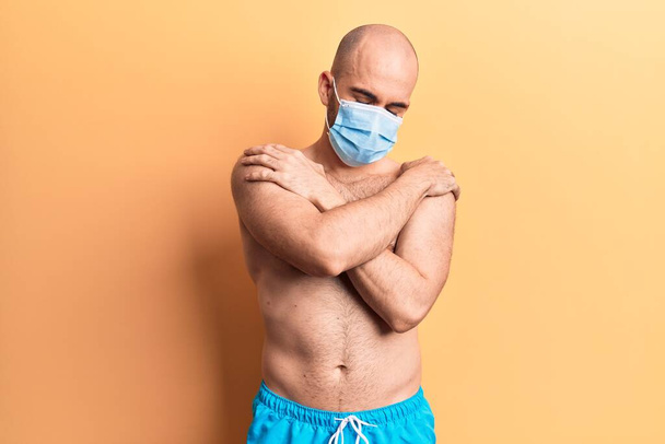 Young handsome bald man shirtless wearing swimwear and medical mask hugging oneself happy and positive, smiling confident. self love and self care  - Фото, изображение