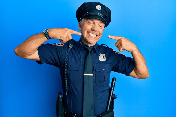 Красивый зрелый мужчина средних лет в полицейской форме, весело улыбающийся и показывающий пальцами зубы и рот. концепция стоматологического здоровья.  - Фото, изображение