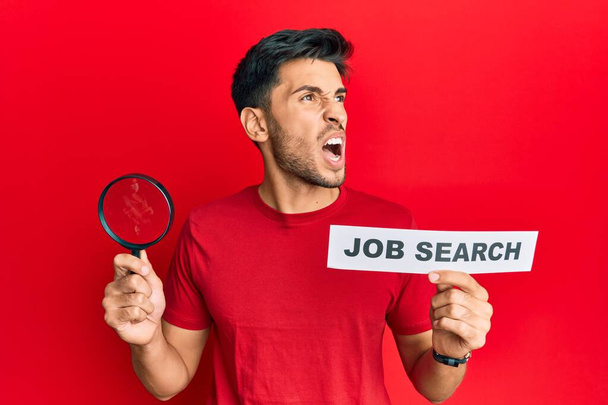 Young handsome man holding magnifying glass for job search angry and mad screaming frustrated and furious, shouting with anger. rage and aggressive concept.  - Photo, Image