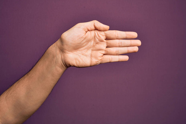 Main de jeune homme caucasien montrant les doigts sur fond violet isolé étirant et atteignant avec la main ouverte pour poignée de main, montrant la paume - Photo, image