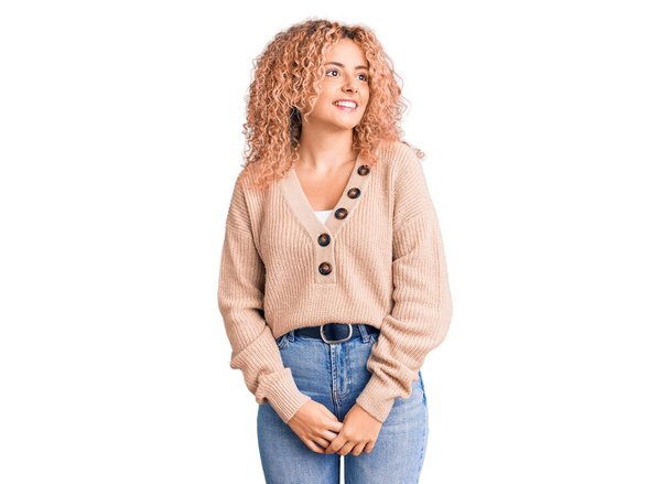 Jeune femme blonde aux cheveux bouclés portant un pull d'hiver décontracté regardant de côté avec le sourire sur le visage, expression naturelle. riant confiant.  - Photo, image