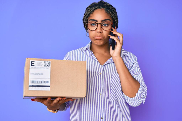 Mujer afroamericana joven con trenzas sosteniendo la caja de entrega llamando a la asistencia deprimida y preocupada por la angustia, llorando enojada y asustada. expresión triste.  - Foto, Imagen