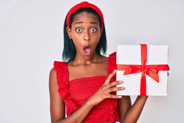 Mujer afroamericana joven sosteniendo regalo asustada y sorprendida con la boca abierta para sorpresa, cara de incredulidad  - Foto, Imagen
