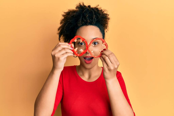 Young african american girl holding red pepper as a glasses afraid and shocked with surprise and amazed expression, fear and excited face.  - Fotografie, Obrázek