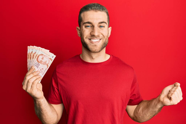Mladý kavkazský muž drží 10 singapurských dolarů bankovky křičí hrdě, slaví vítězství a úspěch velmi vzrušený se zvednutou paží  - Fotografie, Obrázek