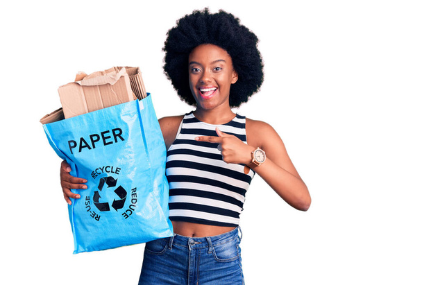 Młoda Afroamerykanka trzymająca worek na recykling z papierem i kartonem uśmiechnięta szczęśliwa wskazując ręką i palcem  - Zdjęcie, obraz